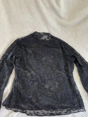Svart spetsblus med lång ärm - Snygg svart blus i genomskinlig spets med blommigt mönster. Blusen har lång ärm och hög hals, perfekt för dig som vill ha en trendig och elegant look. Materialet är tunn spets som ger en cool och stilren vibe.