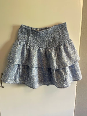Blommig volangkjol från Gina Tricot - Supersöt blå kortkjol från Gina Tricot med små vita blommor och bred smock i midjan. Kjolen har dubbla volanger. 