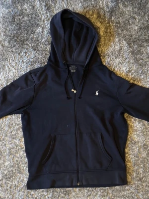 Marinblå hoodie från Polo Ralph Lauren - Snygg marinblå hoodie från Polo Ralph Lauren med dragkedja framtill och klassisk logga broderad på bröstet. Utrustad med huva och justerbara snören samt två fickor framtill. Perfekt för en avslappnad och stilren look.