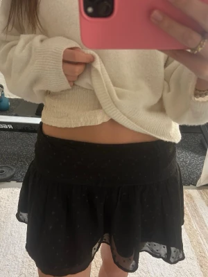 Svart glittrig kortkjol med volang - Supersöt svart kortkjol med volangkant och lätt transparent ytterlager. Kjolen har diskreta prickar i tyget och en bred midjeresår för skön passform. Perfekt att styla med både stickad tröja eller linne och sitter lågmidjade. 