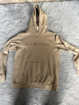 Beige hoodie från Sail Racing - Snygg beige hoodie från Sail Racing med tryckt logga på bröstet. Klassisk känguruficka framtill och huva. Tillverkad i mjukt material, perfekt för en avslappnad och stilren look. Långa ärmar och ribbade muddar vid ärmslut och nederkant.