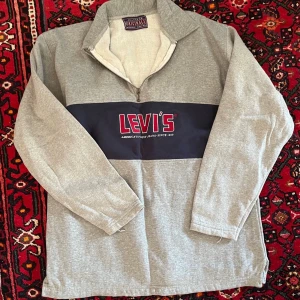 Grå Levi's sweatshirt med dragkedja/quarter zip samt champion byxor - Snygg grå sweatshirt från Levi's med halv dragkedja och hög krage samt grå champion byxor. Stor logga i rött och vitt på mörkblå panel över bröstet. Tillverkad i mjukt bomullsmaterial och har långa ärmar. Perfekt för en avslappnad streetwear-look.