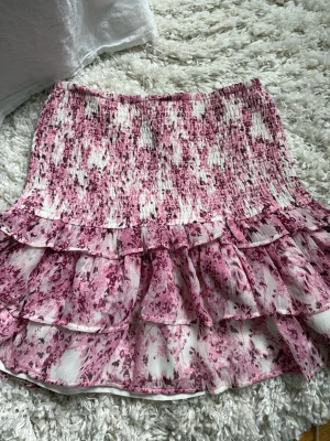 Blommig volangkjol från Gina Tricot - Supersöt kort kjol från Gina Tricot i vitt och rosa med blommigt mönster. Kjolen har smock upptill och dubbla volanglager som ger en flowy känsla. Perfekt för vår och sommar, lätt att matcha med en enkel topp.