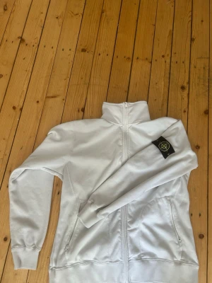 Stone Island - Säljer en clean vit hoodie från Stone Island med hel dragkedja framtill och två fickor med dragkedja. Klassisk patch med logga på vänster ärm. Hoodien har hög krage och är gjord i mjukt bomullsmaterial. Perfekt för en stilren och avslappnad look.