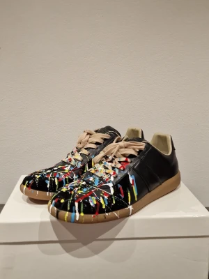 GATS - Unika svarta sneakers med coola färgstänk i rött, blått, gult och vitt. Skorna har beige snörning och en brun sula. De har en klassisk låg profil och är tillverkade i läder med en konstnärlig touch som sticker ut.