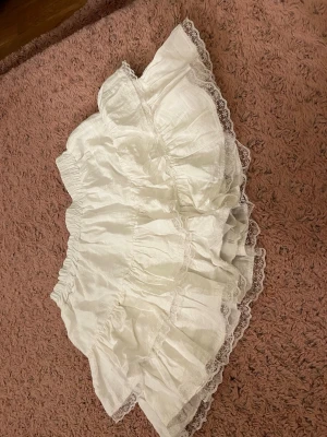Vit volangkjol med spetskant - Söt vit kjol med flera lager volanger och dekorativ spetskant runt om. Kjolen har resår i midjan och är gjord i ett lätt, tunt tyg som ger ett fluffigt intryck. Perfekt för dig som gillar romantisk och feminin stil.