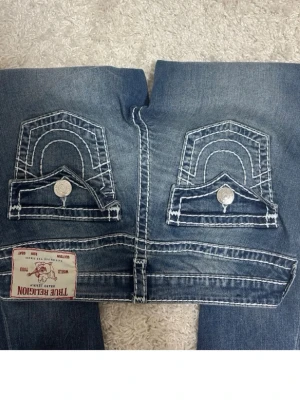 Blå True Religion jeans med kontrastsömmar - Säljer ett par blå jeans från True Religion med tydliga vita kontrastsömmar och stora ficklock bak med silverfärgade knappar. Klassisk femficksmodell och patch med logga bak. Snygg tvättad look och coola detaljer som sticker ut. Ingen defekt o använd typ Max 2 gngr