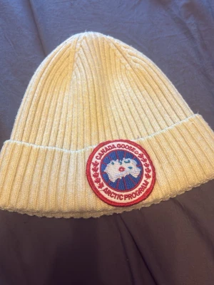 Vit ribbstickad mössa Canada Goose - Säljer en vit ribbstickad mössa från Canada Goose med klassisk rund patch framtill i rött, blått och vitt. Mössan har uppvikt kant och är tillverkad i ett mjukt, värmande material som passar perfekt för kalla dagar.