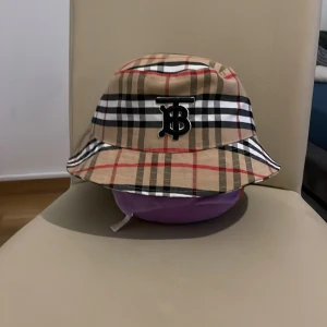 Rutig bucket hat med logotyp ckopia - Snygg bucket hat med klassiskt rutigt mönster i beige, svart, vit och röd. Hatten har en broderad logotyp framtill och är perfekt för en trendig look.