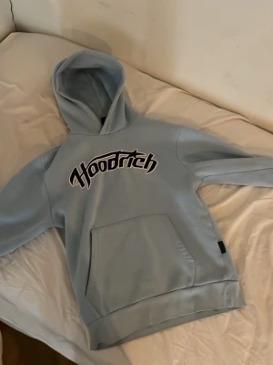Ljusblå hoodie med Hoodrich-tryck - Snygg ljusblå hoodie med stor magficka och svartvitt Hoodrich-tryck på bröstet. Klassisk huva och relaxed fit, perfekt för en chill och street-inspirerad stil. Tillverkad i mjukt material som känns skönt mot huden.