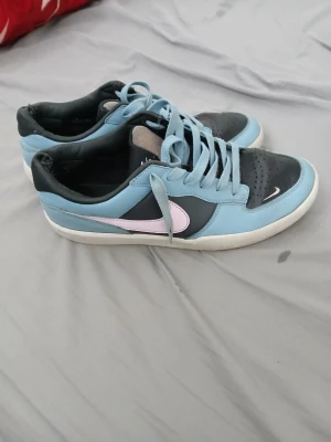 Nike blå och svarta sneakers - Snygga sneakers från Nike i blått och svart med ljusrosa swoosh. Skorna har lågt skaft, klassisk snörning och vit sula. Ovandelen är i en mix av läder och textil, vilket ger en sportig och fräsch look. Skorna är lite sönder där bak men man märker inte. Nypris är 1250 min pris är 300