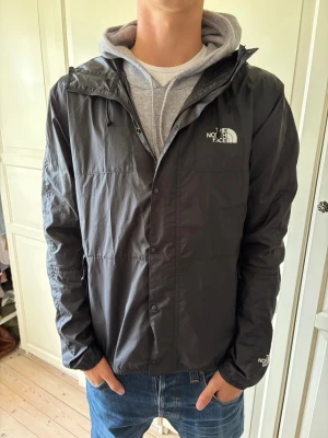Svart vindjacka från The North Face - Snygg svart vindjacka från The North Face med huva och dragkedja. Jackan har logga på bröstet och ärmen, samt justerbar huva. Tillverkad i lätt och tåligt material, perfekt för blåsiga dagar. Enkel och stilren design som passar till det mesta. Jackan är i storlek L. Hör av er vid frågor och priset är inte hugget i sten!