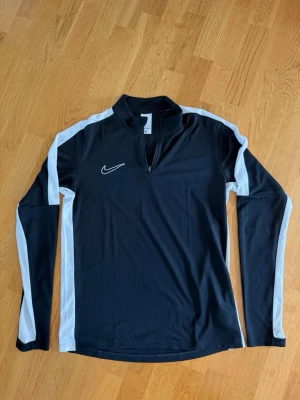 Svart långärmad tröja från Nike - Svart långärmad träningströja från Nike med vita detaljer längs ärmarna och axlarna. Tröjan har halv dragkedja framtill och är tillverkad i ett lätt, stretchigt material som passar perfekt för träning och sportaktiviteter.