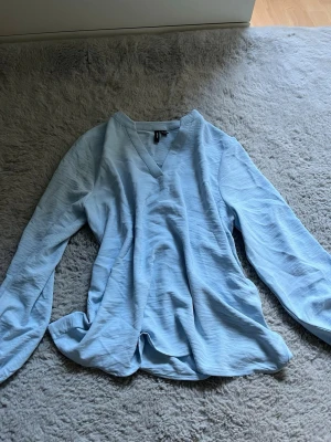 Ljusblå blus från Vero Moda - Snygg ljusblå blus från Vero Moda med v-ringning och långa ärmar. Tillverkad i ett lätt och mjukt tyg som ger en avslappnad look. Perfekt att styla med jeans eller kjol för en fräsch outfit.