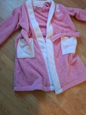 Rosa morgonrock Liverpool FC för barn till 5-6 år (går ålder) - Mysig rosa morgonrock från Liverpool FC med vita detaljer, två stora fickor och broderad logga på bröstet. Tillverkad i fluffigt, mjukt material och har knytskärp i midjan. Perfekt för att chilla hemma. Rätt så fint skick använt när jag va yngre några gånger på stranden.