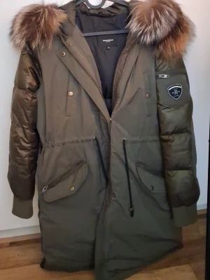 Rockandblue jacka  - Snygg olivgrön med stor huva och fluffig päls i bruna toner. Jackan har dragkedja, knappar, snörning i midjan och flera praktiska fickor. Ribbade muddar vid ärmslut och patch på ärmen ger en cool look. Perfekt för kalla dagar. Jackan är I mycket bra skick dock har dragkedjan gått av,men den följer med så man kan laga själv eller hos skräddare. St 38