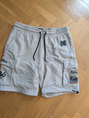 Beigea cargo shorts med Kodak-tryck - Säljer ett par beigea cargo shorts med coola Kodak-print och färgglada detaljer. Shortsen har stora fickor på sidorna, dragsko i midjan och justerbar nederkant. Perfekta för en avslappnad och trendig look.
