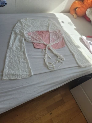 Vit spetsbolero och rosa linnetopp - Säljer en söt topp med rosa linnetopp och en vit, långärmad bolero i spets. Boleron har knytband framtill och genomskinligt spetsmönster. Perfekt för dig som vill ha en romantisk och trendig look.