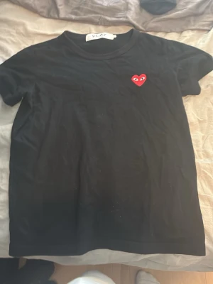 Svart Comme des Garçons PLAY t-shirt - Svart t-shirt från Comme des Garçons PLAY med det ikoniska röda hjärtat med ögon broderat på bröstet. Klassisk rund hals och korta ärmar. Perfekt basic med en twist för dig som gillar streetwear och stilrena plagg.