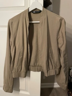 Beige bomberjacka från Zara - Stilren beige bomberjacka från Zara med dragkedja framtill och två fickor. Jackan har ribbade muddar vid ärmslut och midja för en snygg passform. Perfekt att slänga över en t-shirt eller hoodie för en avslappnad look.