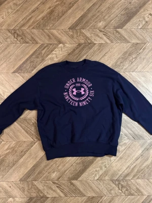 Mörkblå sweatshirt Under Armour - Mörkblå sweatshirt från Under Armour med rund halsringning och tryck i rosa framtill. Tröjan har ribbade muddar vid ärmslut och nederkant samt en avslappnad passform. Perfekt för en sportig och casual stil.