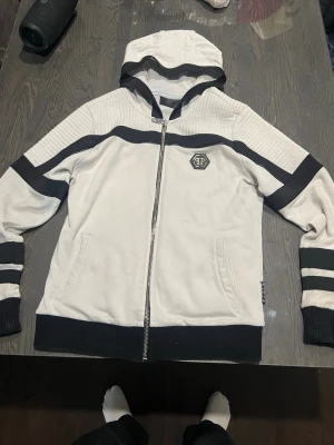 Vit hoodie från Philipp Plein - Snygg vit hoodie från Philipp Plein med svarta detaljer och dragkedja framtill. Hoodien har svarta ränder på ärmarna, ribbade muddar och en logga på bröstet. Tillverkad i mjukt material och har två fickor framtill samt huva.