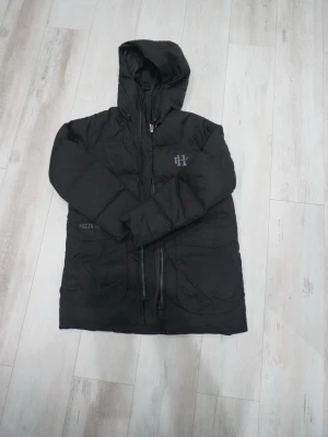Svart dunjacka från Felix Hardy - Snygg svart dunjacka från Felix Hardy med huva och dragkedja framtill. Jackan har diskret logga på bröstet och är tillverkad i ett glansigt material som ger en modern look. Perfekt för kalla vinterdagar när du vill hålla dig varm och stilren.