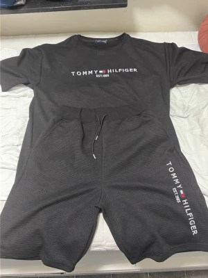 Mörkgrått set från Tommy Hilfiger - Snyggt svart set från Tommy Hilfiger med t-shirt och shorts. Båda delarna har logga och texten 'EST. 1985' tryckt framtill. Shortsen har dragsko i midjan och sidofickor. Perfekt för en avslappnad och trendig look.