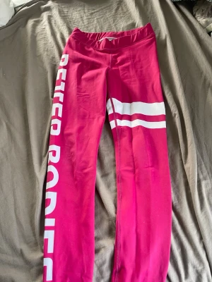 Rosa leggings med vit text - Snygga rosa leggings med vit text längs ena benet och två vita ränder på låret. Perfekt för dig som gillar sportiga och trendiga plagg. Stretchigt material som sitter bekvämt och följer kroppens rörelser.