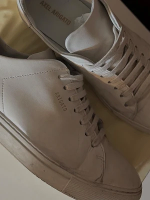 Vita sneakers från Axel Arigato - Stilrena vita sneakers från Axel Arigato i slät läder med diskret logga på sidan. Klassisk låg modell med vita snören och minimalistisk design som passar till det mesta. Perfekta för dig som gillar clean och modern stil. Skorna är i okej skick och är endast smutsiga, vänstra skon har dock en skada som visas på andra bilden. Skriv gärna privat för mer bilder eller frågor!