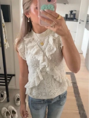 Vit stickad topp med volanger - Supersöt vit stickad topp med volangdetaljer framtill och på axlarna. Toppen är ärmlös och har en rund halsringning. Perfekt att styla med jeans eller kjol för en trendig look.