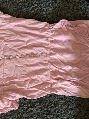 Ljusrosa kort klänning med knappar - Söt ljusrosa klänning i kort modell med dekorativa knappar framtill. Klänningen har korta ärmar och är tillverkad i ett mjukt tyg, perfekt för varma dagar. Enkel och stilren design som passar till många tillfällen. SAKNAS EN KNAPP!!