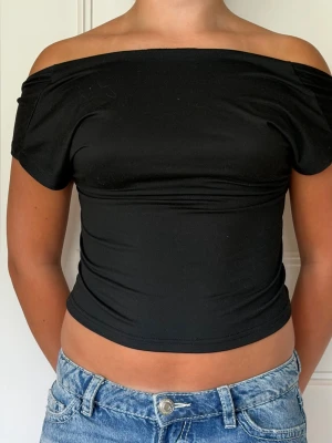 Svart offshoulder topp med öppen rygg - Snygg svart offshoulder topp med korta ärmar och helt öppen rygg. Toppen är croppad och har en tight passform som framhäver figuren. Perfekt för dig som vill ha en trendig och stilren look.