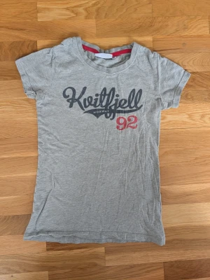 Grå Kvitfjell t-shirt med tryck - Snygg grå t-shirt med texten 'Kvitfjell Norway 92' tryckt framtill i svart och rött. Klassisk rund halsringning och korta ärmar. Perfekt för en avslappnad stil och enkel att matcha med jeans eller shorts.