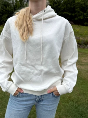Hoodie med huva , gräddvit - Säljer en enkel och gräddvit hoodie med huva och dragsko. Hoodien har en avslappnad passform, långa ärmar och ribbade muddar vid ärmslut och nederkant. Perfekt till jeans för en chill look. Flossad insida, varm och go. Sparsamt använd, så fint skick. Några små noppor finns men tas lätt bort ned noppmaskin. Hel och ren och inga bitmärken på snörena. 