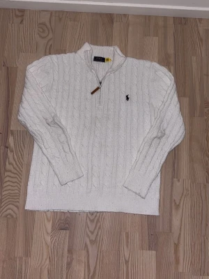 Vit kabelstickad tröja Polo Ralph Lauren - Kabelstickad vit tröja från Polo Ralph Lauren med halv dragkedja och klassisk broderad logga på bröstet. Tröjan har hög krage och ribbade muddar. Perfekt för lager på lager och en clean look.