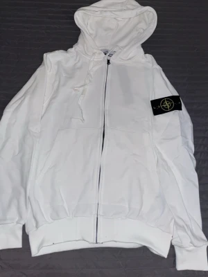 Vit hoodie med dragkedja och patch - Snygg vit hoodie med dragkedja framtill, stor huva och fickor. På vänster ärm sitter en svart patch med gult och grönt broderat märke. Hoodien har ribbade muddar och en avslappnad passform. Perfekt för en clean och stilren look.