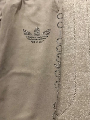 Beige mjukisbyxor med strassdetaljer ,Ej äkta - Snygga beige mjukisbyxor från Adidas med glittrande strassdetaljer längs benet och loggan framtill. Byxorna har resår i midjan, fickor och muddar vid bensluten. Materialet är mjukt och bekvämt, perfekt för chill dagar.