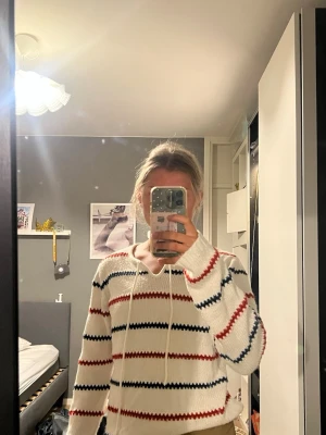 Vit stickad tröja med ränder - Vit stickad tröja med röda och marinblå horisontella ränder. Tröjan har en avslappnad passform, långa ärmar och en huva med dragsko. Perfekt för en chill och trendig look.