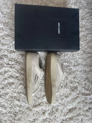 Beige Saint Laurent sneakers - Snygga beige sneakers från Saint Laurent med lågt skaft och klassisk siluett. Skorna har texten 'Saint Laurent' på sidan, beige snörning och ovandel i canvas. Perfekta för dig som gillar stilrena och exklusiva sneakers.