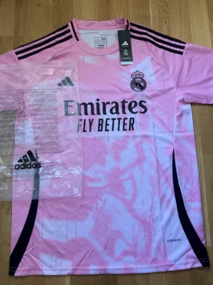 Rosa Real Madrid matchtröja Adidas - Säljer en rosa Real Madrid matchtröja från Adidas med svart logga och detaljer. Tröjan har korta ärmar, rund hals och svart/vita ränder på axlarna. Materialet är lätt och andas, perfekt för träning eller match. Emirates Fly Better-tryck på bröstet.