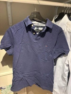 Blå pikétröja från Tommy Hilfiger - Snygg blå pikétröja från Tommy Hilfiger med klassisk krage och två knappar framtill. Tröjan har korta ärmar och den ikoniska lilla loggan broderad på bröstet. Perfekt till jeans eller shorts för en avslappnad stil. Storleken är i Amerikanskt mått, men den passar som Medium