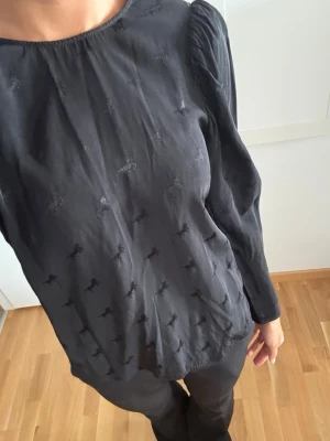 Svart blus med puffärm och diskret mönster - Snygg svart blus med puffärm, små diskreta hästar och rund halsringning. Blusen har ett diskret ton-i-ton broderat mönster över hela tyget och är tillverkad i ett tunt, mjukt material som ger ett elegant fall. Perfekt att styla med både jeans och kostymbyxor.
