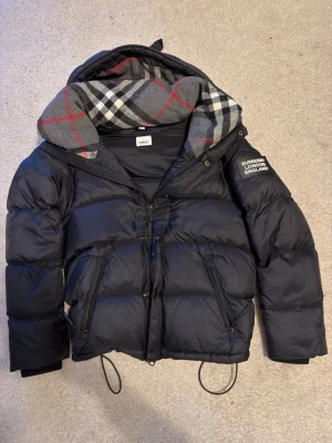 Svart pufferjacka från Burberry - Säljer en svart pufferjacka från Burberry med huva och klassiskt rutigt foder i grått, vitt, rött och svart. Jackan har dragkedja, snörning i nederkant och en patch på ärmen med texten 'Burberry London England'. Perfekt för kalla vinterdagar.