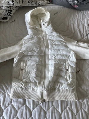 Vit pufferjacka med huva - Snygg vit pufferjacka med huva och dragkedja framtill. Jackan har ribbade muddar och ärmar i stickat material samt två fickor med knappar. Puffig design och stilren look, perfekt för kalla dagar.