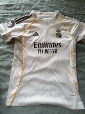 Real Madrid matchtröja Mbappé #9 - Säljer en vit Real Madrid matchtröja från Adidas med Mbappé och nummer 9 på ryggen. Tröjan har svarta detaljer, gula sömmar och klubbmärke på bröstet. Tillverkad i lätt funktionsmaterial (Aeroready) och har sponsortryck på framsidan.