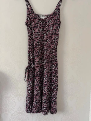 Sommarfin klänning - Sommarfin klänning, luftig med band i midjan som går strax ovan knäna. I botten svart/mörkblå med vita/lila/rosa blommor. 95% viskos och 5% elastan.