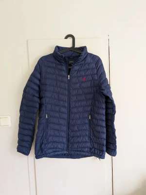 Polo Ralph Lauren quiltad jacka marinblå XL (18–20) - Snygg och klassisk quiltad jacka från Polo Ralph Lauren i marinblått 💙 Röd broderad Polo-logga på bröstet och två fickor framtill med dragkedja. Storlek XL (18–20), junior. Lätt vadderad och perfekt som vår-/höstjacka eller under mildare vinterdagar. • Marinblå • Röd broderad Ralph Lauren-logga • Dragkedja framtill • Två fickor med dragkedja • Yttertyg: 100% polyester • Foder: 100% nylon • Fyllning: 100% polyester • Fint begagnat skick – se bilder för skick Säljes då den inte längre används.