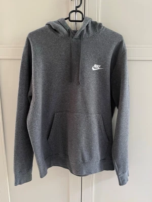 Mörkgrå Nike hoodie med huva - Snygg mörkgrå hoodie från Nike med klassisk logga i vitt på bröstet. Hoodien har en stor känguruficka framtill, justerbar dragsko i huvan och ribbade muddar vid ärmar och nederkant. Se annons med tillhörande matchande byxor för att köpa helt set. 