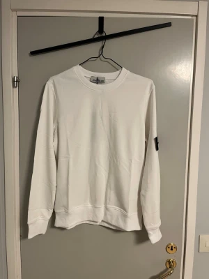 Vit tröja Stone Island - Säljer en stilren vit sweatshirt från Stone Island med klassisk rund hals och ribbade muddar. Tröjan har den ikoniska svarta Stone Island-loggan på vänster ärm som går att ta av. Perfekt för dig som gillar enkel och clean stil.